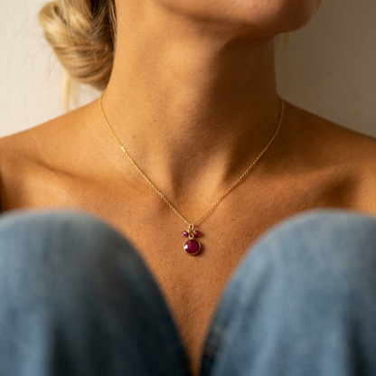 A close-up lifestyle shot of a model wearing a delicate gold chain necklace with a small ruby circle pendant featuring two ruby gemstones, framed by the model's neck and blue jeans.